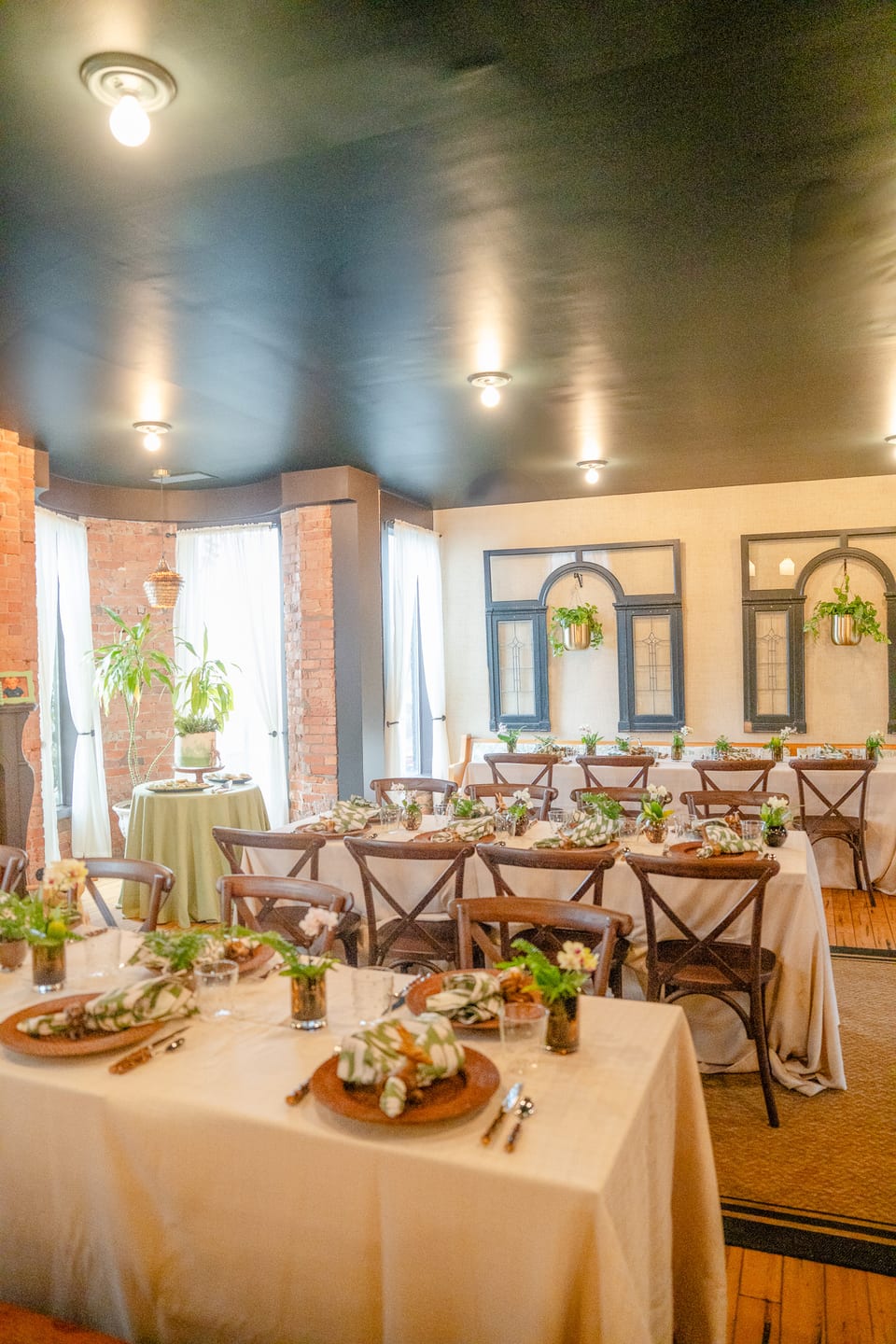 Long banquet tables set with cross-back chairs, neutral linens, and small floral arrangements in The Alexander's Turret Room — brick columns and ivory drapes framing the bay window, navy ceiling overhead.