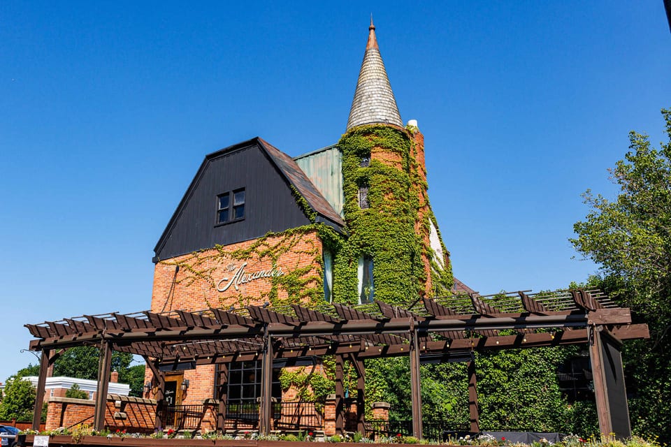 The Alexander — red brick Victorian mansion with ivy climbing the turret, cone-topped roof, script signage, and the pergola out front against a clear blue sky