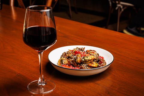 A glass of red wine on a walnut table beside a bowl of crispy brussels sprouts