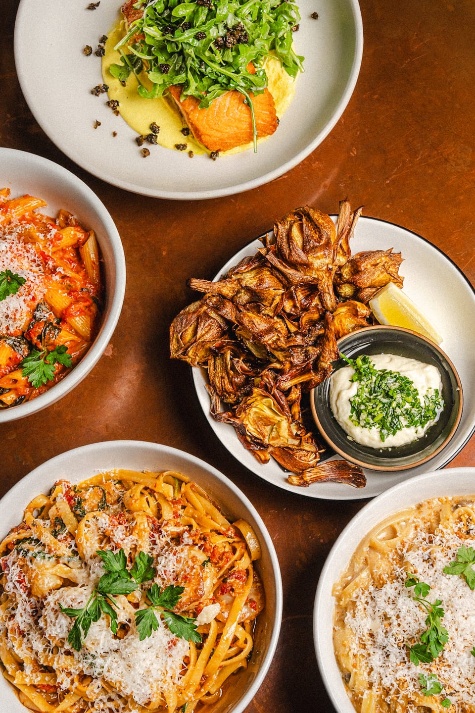 Overhead spread of catering dishes — fried artichokes with whipped ricotta, salmon over saffron sauce with arugula, rigatoni in tomato, and two creamy pastas plated across a warm copper table
