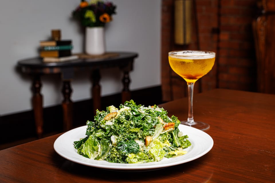 Kale caesar salad plated beside an amber craft cocktail in a coupe glass — dark wood table, soft background of books and a vase of sunflowers