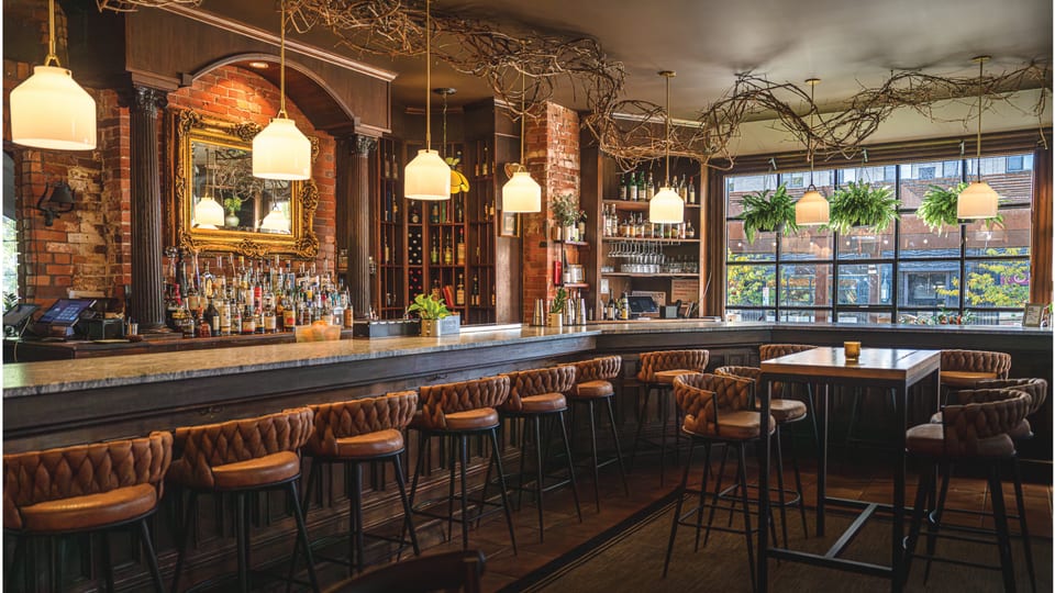 The Alexander bar — brass pendant lights, leather-back stools, brick wall, evening