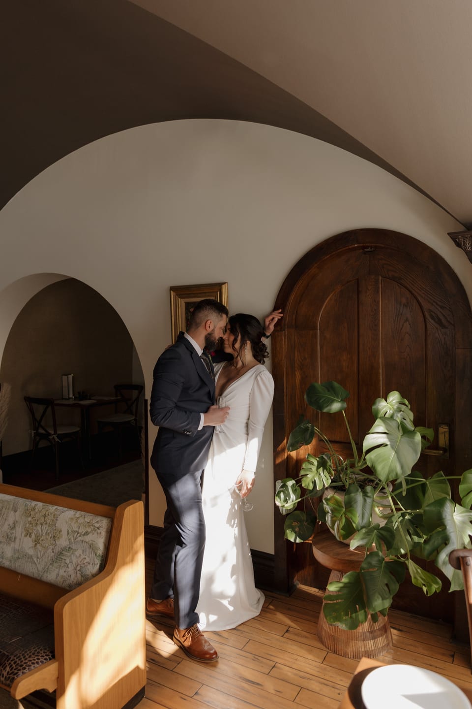 Couple kissing under the curved white archway with monstera plant in foreground