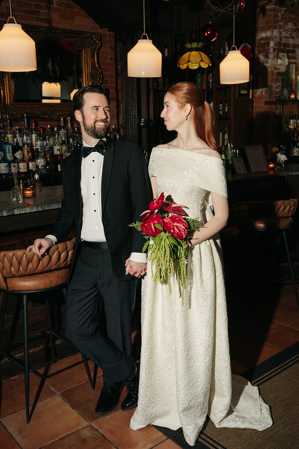 Bride and groom standing at the bar holding a red anthurium bouquet